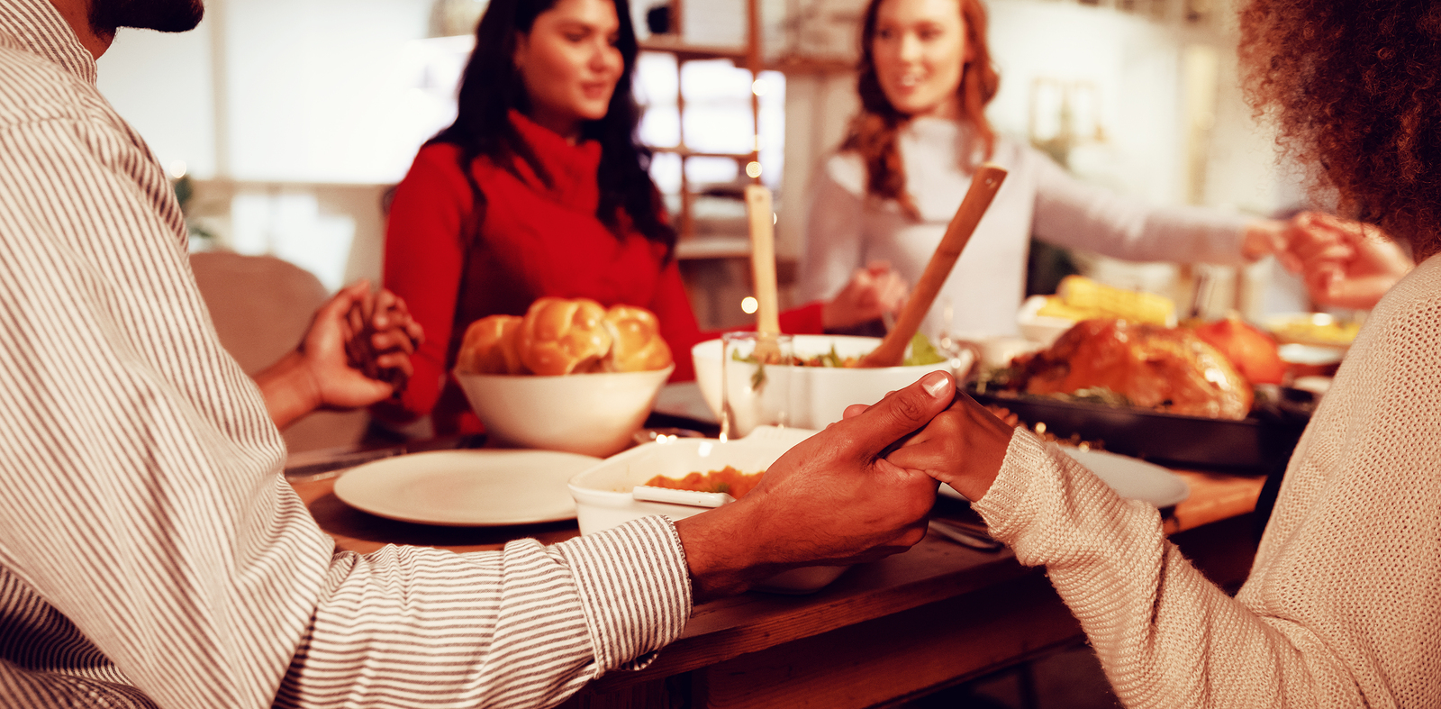 Over the shoulder view of a group of young adult multi-ethnic male and female friends sitting around a table holding hands saying grace before eating thanksgiving dinner at home together