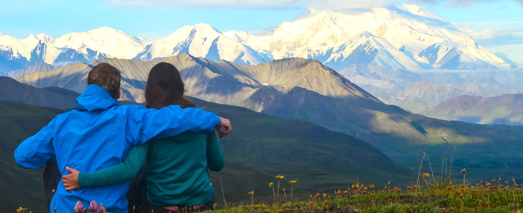 A couple sits with arms around shoulders backs to camera view of mountains in the distance